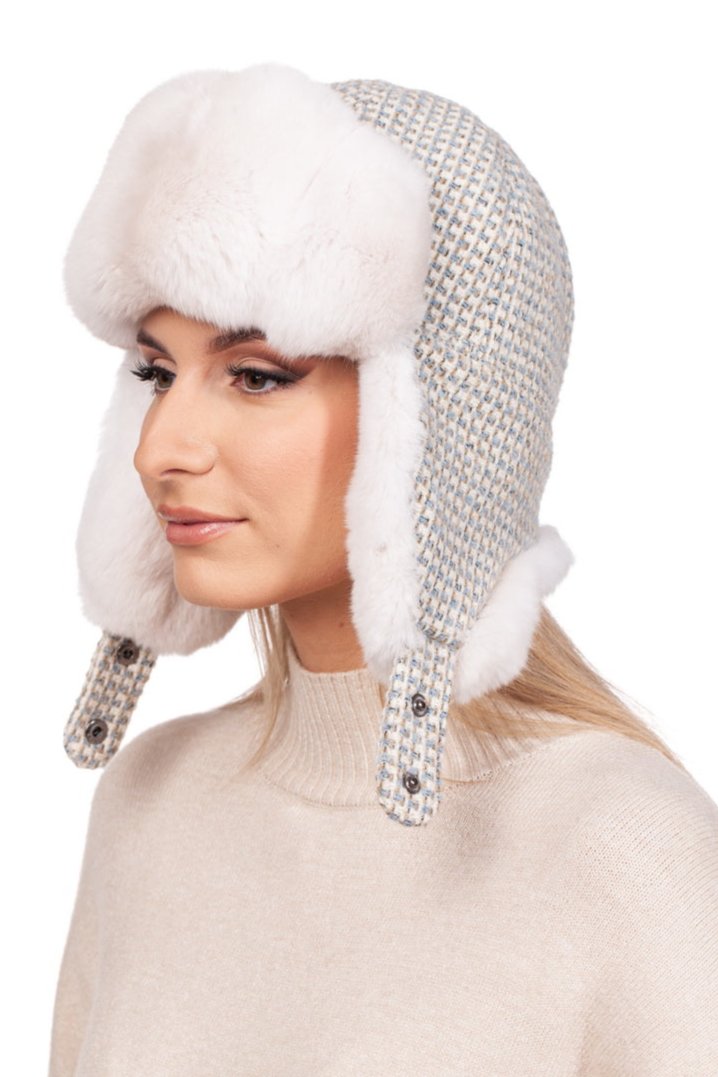 The individual is adorned in a luxurious Cream White Chinchilla Fur Ushanka Hat by FurbySD, complete with ear flaps and buttons, and boasting a cozy fur-like lining. They also wear a beige sweater while set against a plain white background.