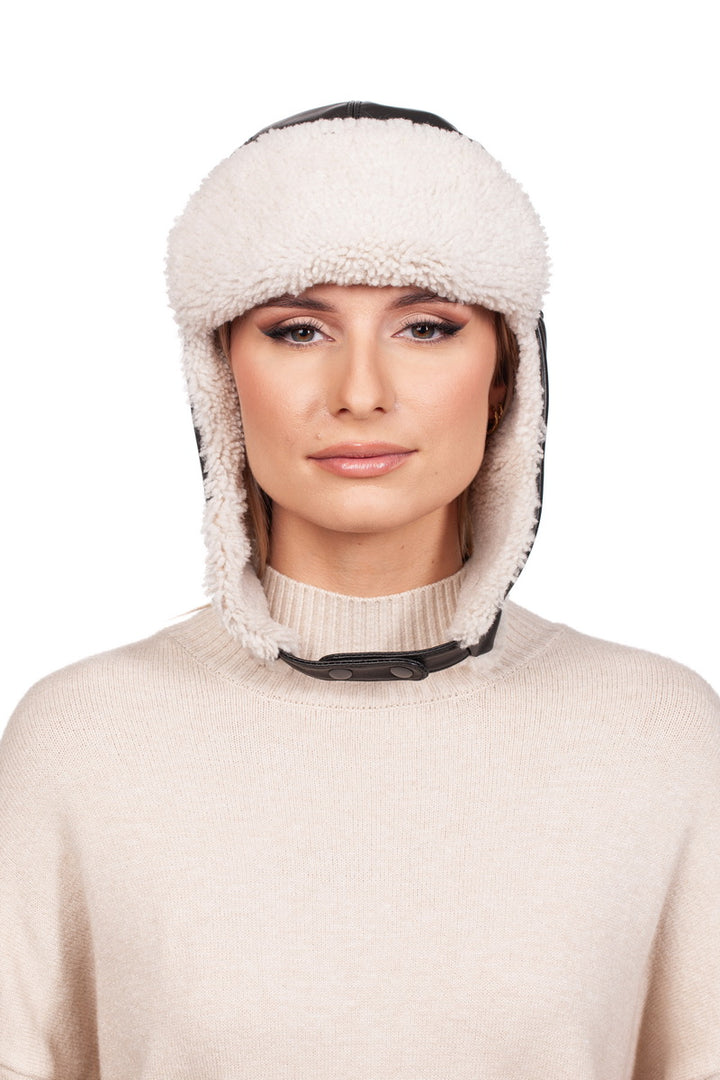 A person is wearing a beige sweater and the White Shearling Fur Aviator Bomber Hat by FurbySD, which features fluffy white shearling fur trim. The hat covers their ears and includes a black strap under the chin, all set against a plain white background.