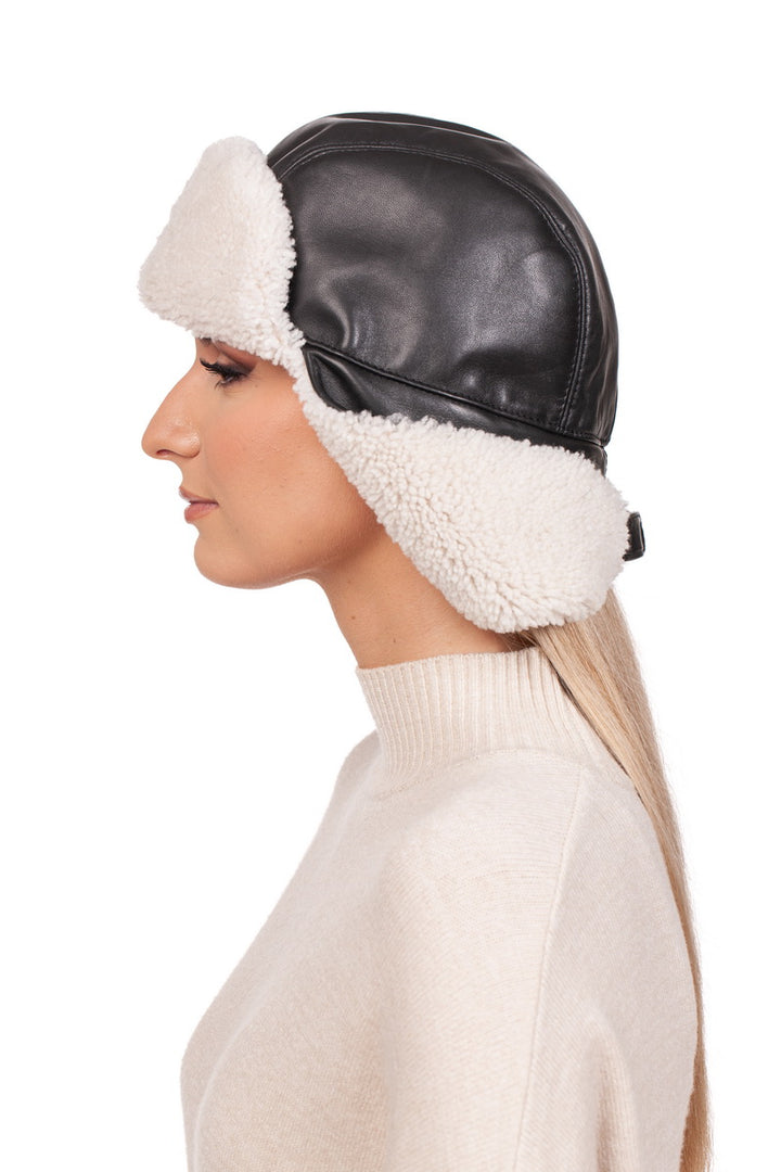 From a side angle, an individual is seen wearing the White Shearling Fur Aviator Bomber Hat by FurbySD, crafted from black Nappa leather and lined with white fleece. To complement the hat, they pair it with a light beige turtleneck sweater, set against a plain white background.