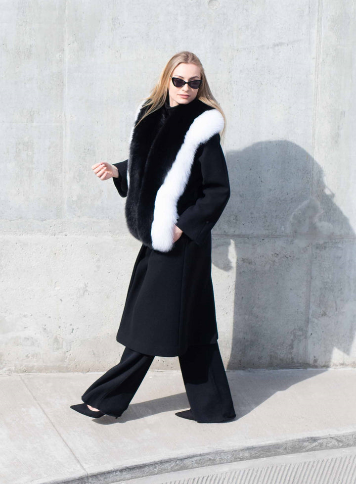 A person wearing a long black coat accessorized with the luxurious Black And White Fox Fur Stole by FurbySD, along with black pants and sunglasses, stands against a concrete wall. They keep their hands in their pockets while gazing to the side.