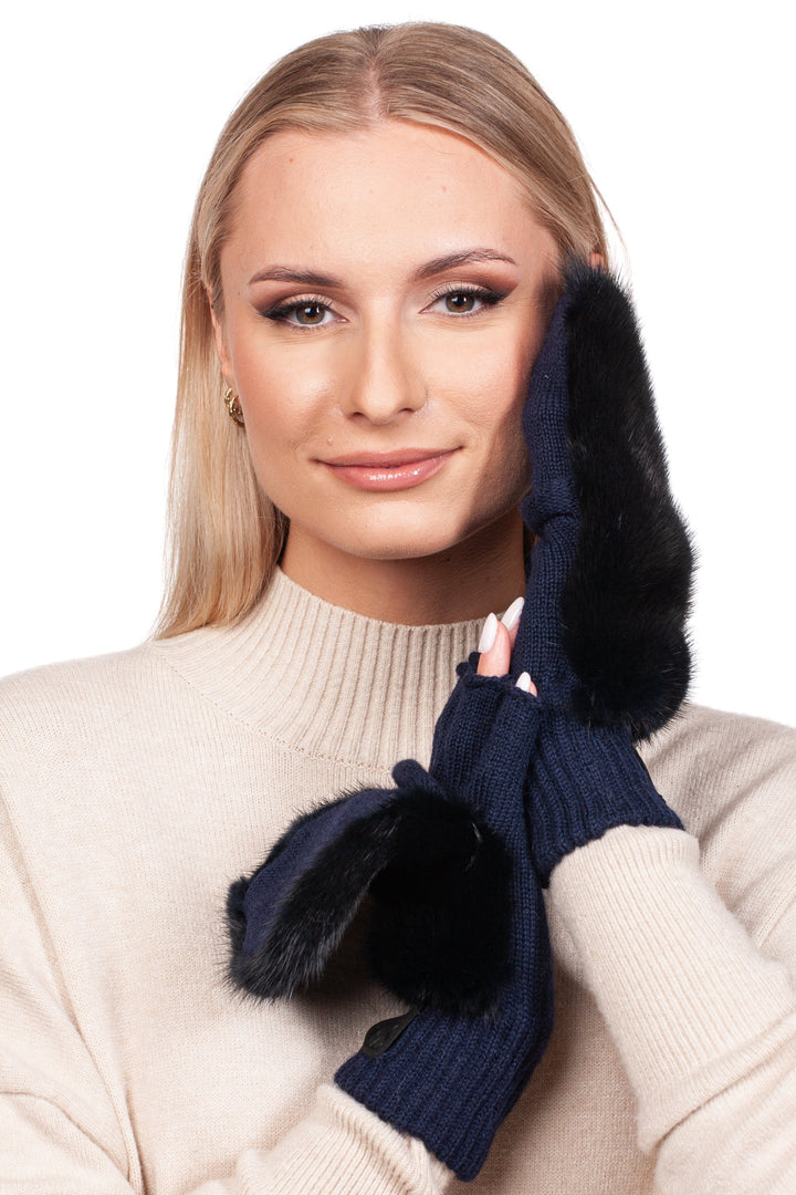 A person with long hair wears a beige sweater and smiles while holding their gloved hands to their face. The FurbySD Dark Blue Mink Fur Flip Top Mittens, which are navy blue with black fur trim and open at the fingers, complete the look against a white background.