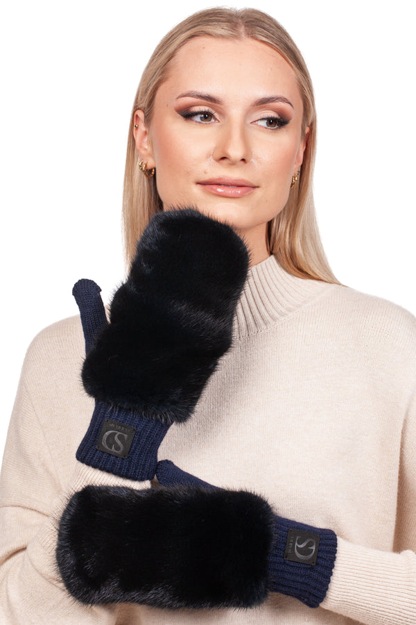 A person with long blonde hair is wearing cozy handcrafted luxury Dark Blue Mink Fur Flip Top Mittens from FurbySD and a cream-colored sweater. They are gazing off to the side with a gentle expression, holding one mitten to their chin.