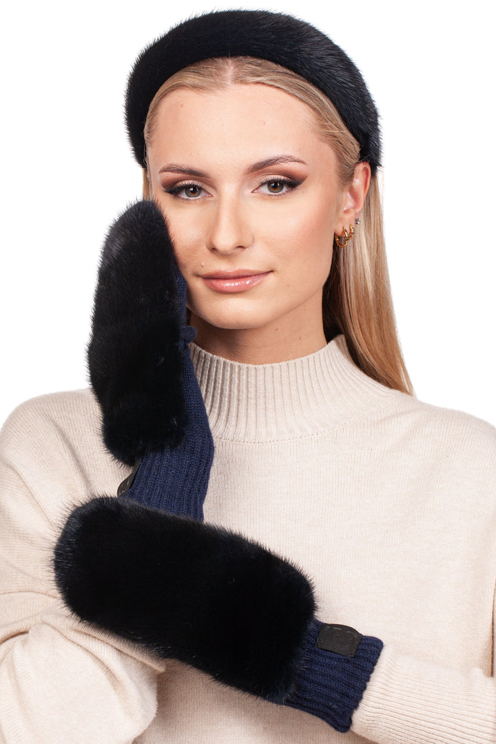 A woman with long blonde hair wearing a beige sweater showcases a luxury winter accessories set from FurbySD, featuring dark blue mink fur mittens and a matching headband. Posing with one hand gently touching her cheek, her expression is calm and she wears subtle makeup complemented by hoop earrings against a white background.