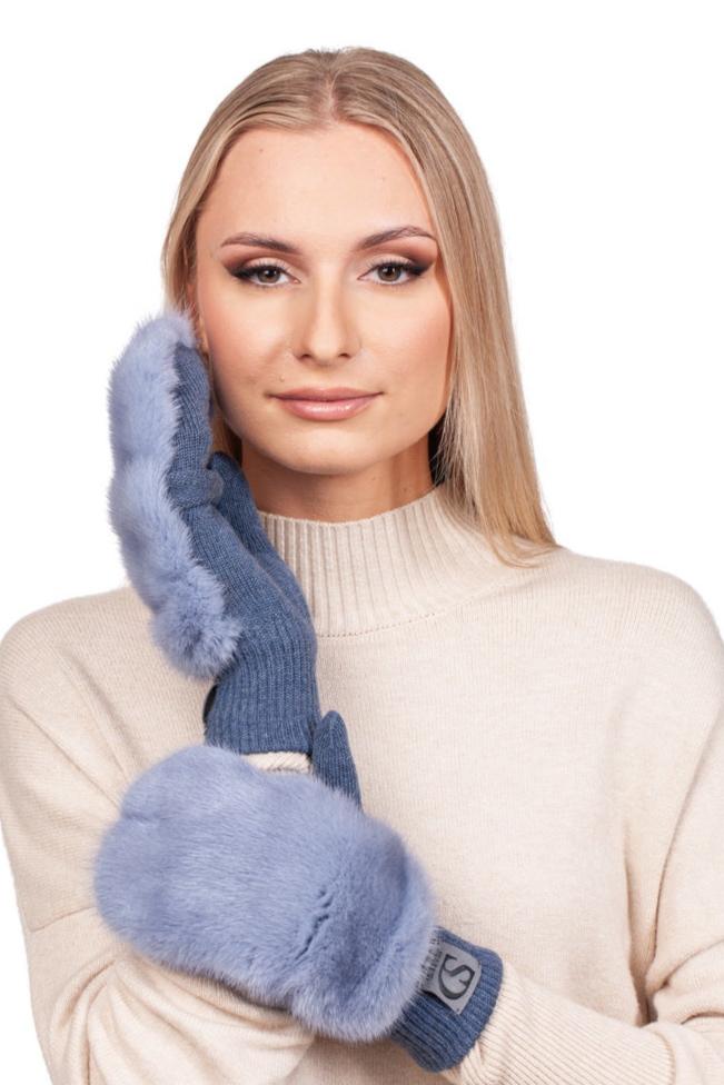 A woman with long blonde hair wears a beige turtleneck sweater, delicately holding a luxurious pair of Denim Blue Mink Fur Flip Top Mittens by FurbySD to her cheek. She smiles softly at the camera, exuding warmth and elegance in her choice of winter accessory.