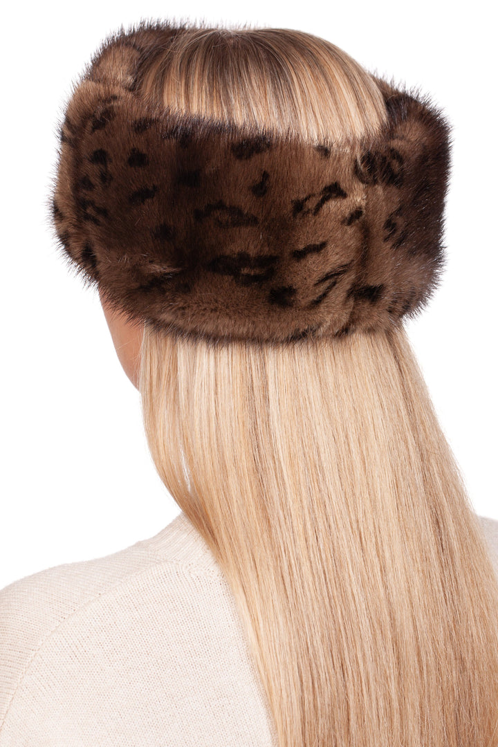 A person with long, straight blonde hair wears a luxurious winter accessory from FurbySD: the Leopard Print Mink Fur Headband in brown, covering their ears. The individual is turned away, displaying the back of their head while wearing a cream-colored top featuring textured detail on the shoulder.