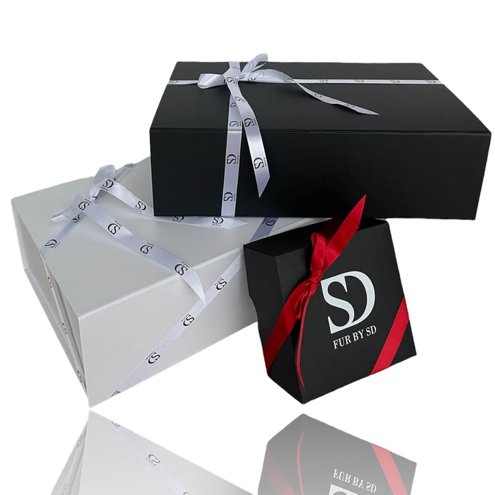 Three gift boxes stacked atop each other: a flat black box with a gray ribbon on top, a larger white box with a similar gray ribbon in the middle, and a smaller black box with a red ribbon at the bottom, featuring "FurbySD" text and logo. Perfect for gifting winter accessories like the Black Sheepskin Hat With a Brim.