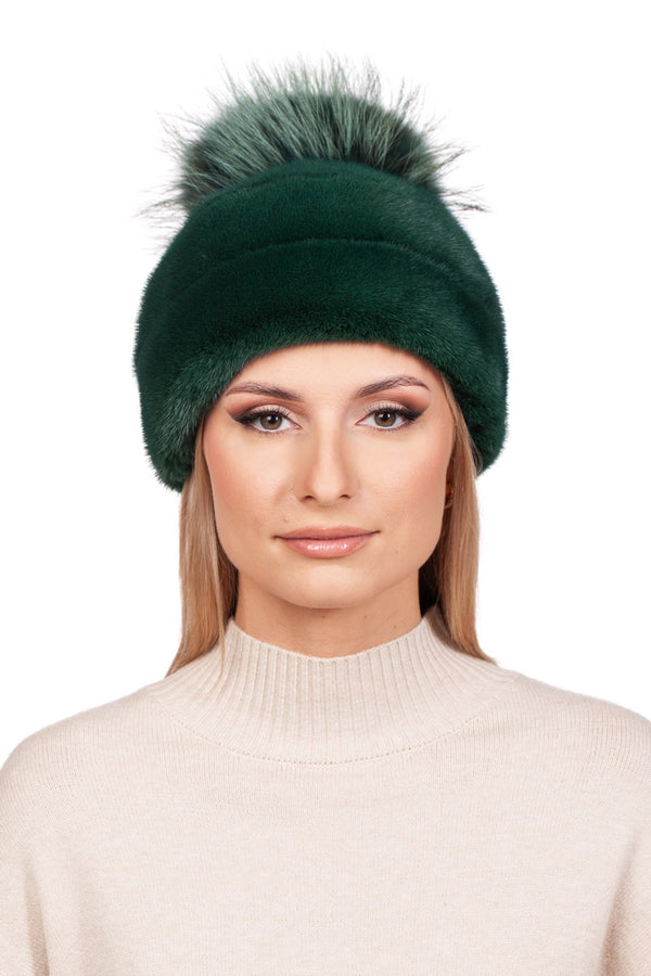 A person with straight, light brown hair is wearing a cozy FurbySD Green Mink Fur Beanie Hat With Fox Fur Bobble. They are dressed in a beige, high-necked sweater against a plain white background.