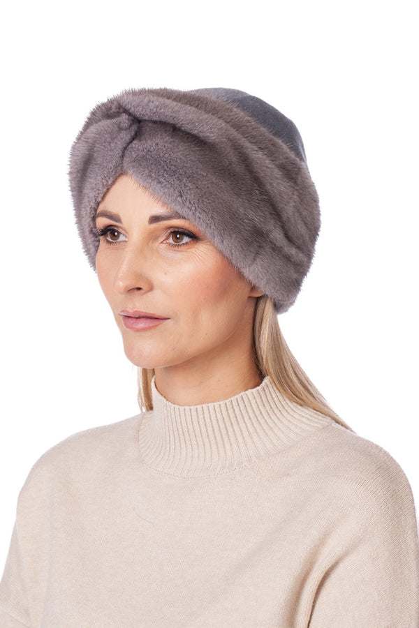 A woman with light skin and long blonde hair wears a soft, gray Sapphire Mink Fur Turban Hat by FurbySD paired with a light beige turtleneck sweater. She is looking slightly to the side, exuding a calm and relaxed expression against a plain white background.