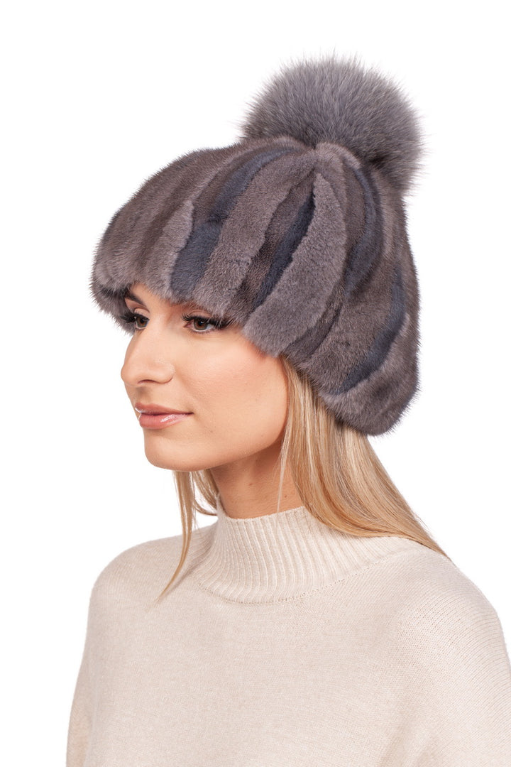 A woman wears the Patched Grey Mink Fur Hat with Fox Bobble from FurbySD, showcasing its handcrafted design with multi-toned stripes. She pairs it with a cream-colored turtleneck sweater and lets her long blonde hair flow freely.