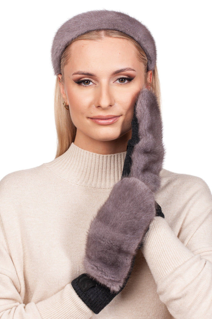 A person wearing a beige sweater, elegantly styled with luxury winter accessories, showcases a FurbySD Grey Mink Fur Headband and Mittens. They gently touch their cheek with the mittened hand while gazing at the camera.