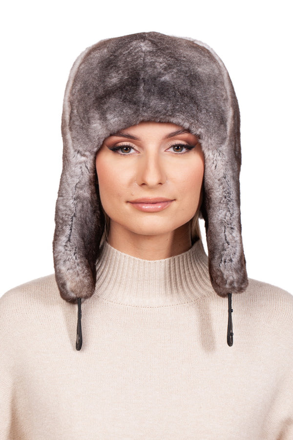 A person with a neutral expression is wearing a FurbySD Grey Rex Rabbit Fur Trapper Hat, complete with ear flaps that hang down. The hat features a soft, gray texture and the individual is dressed in a beige turtleneck sweater. The background is plain white.