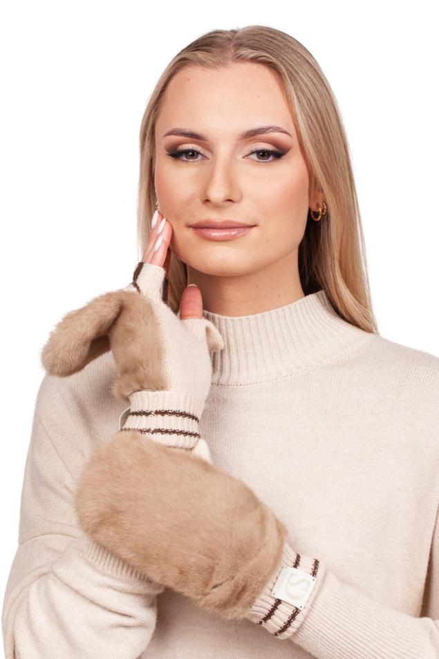 A woman with long blonde hair is wearing a beige sweater and FurbySD's Beige Flip Top Mink Fur Mittens, featuring a playful ear design. Her gentle smile complements her thoughtful pose, while the mittens boast a striped pattern around the wrists.