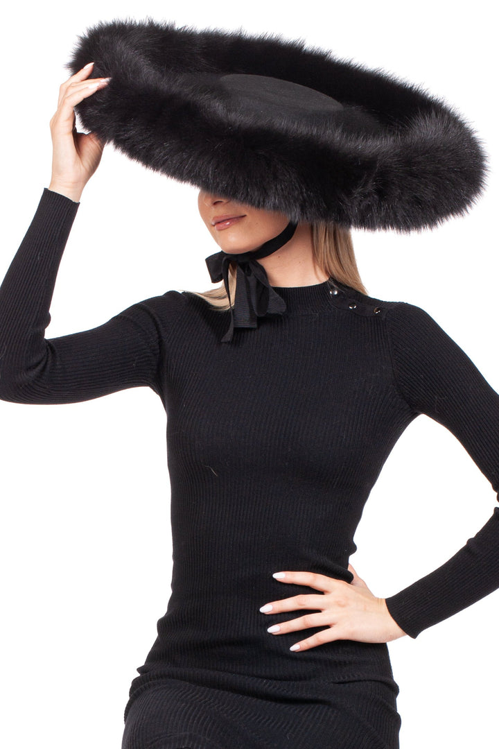 A fashion model dressed in a black long-sleeve dress poses with one hand on her hip. She is holding a FurbySD Black Fox Fur Trimmed Wide Brim Fedora Hat above her head, its fluffy edges partially obscuring her face, set against a white background.