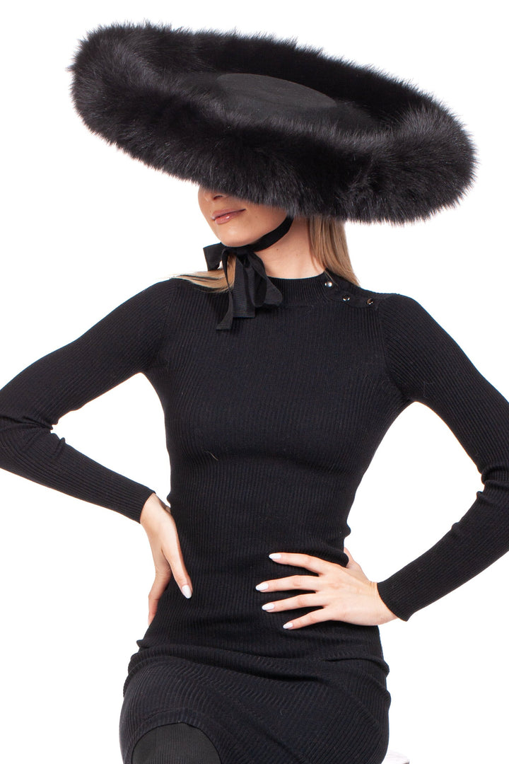 A woman in a fitted black dress poses with one hand on her hip, wearing the FurbySD Black Fox Fur Trimmed Wide Brim Fedora Hat, which features an oversized design with a wide, fluffy fox fur trim that obscures her eyes. The hat is secured with a ribbon under her chin against the backdrop of a plain white background.