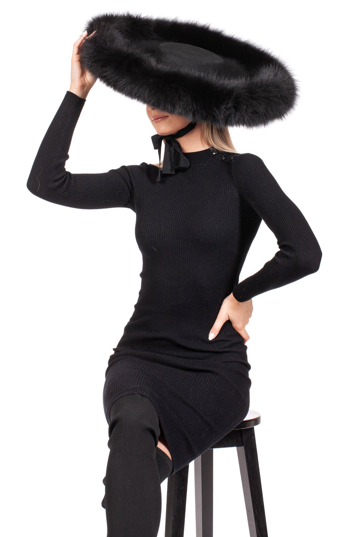 A person sits on a stool wearing a black dress and a FurbySD Black Fox Fur Trimmed Wide Brim Fedora Hat, obscuring their face. Their right hand rests on the stool while the left hand adjusts the hat. The outfit is elegant and monochromatic.