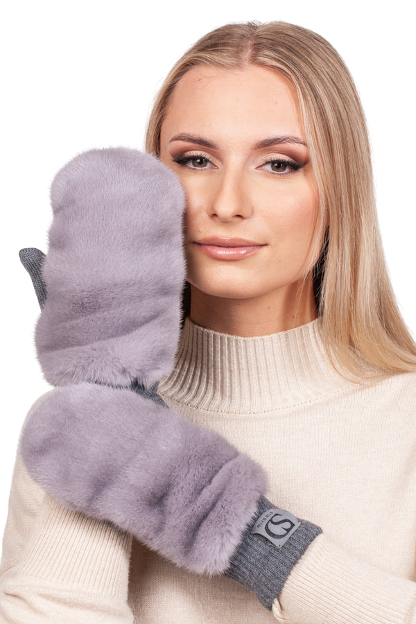 A person with long blonde hair, dressed in a cream sweater, holds a FurbySD Lavender Mink Fur Flip Top Mitten gently against their cheek. With a calm expression, they wear the other luxury mitten on their hand, which is lifted close to their face.