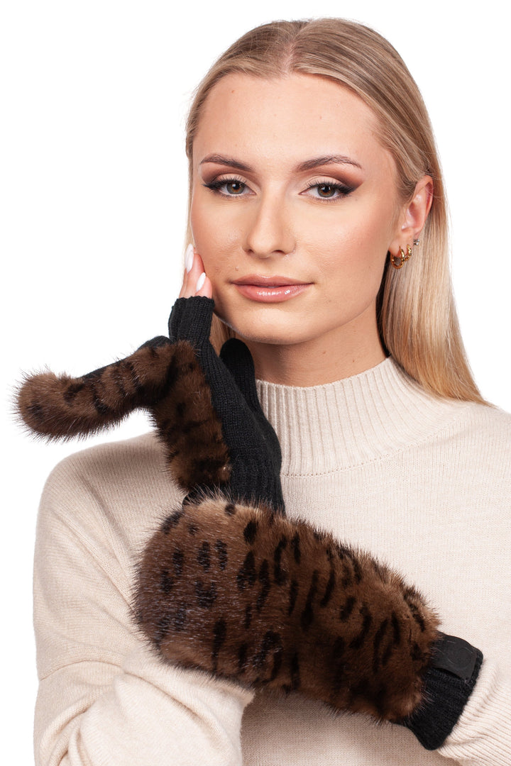 A woman with blonde hair is wearing a beige sweater and a pair of Leopard Print Mink Fur Flip Top Mittens by FurbySD. The mittens feature black fabric adorned with sections of brown mink fur, creating a leopard print look. She is touching her cheek with one hand and gazing at the camera.