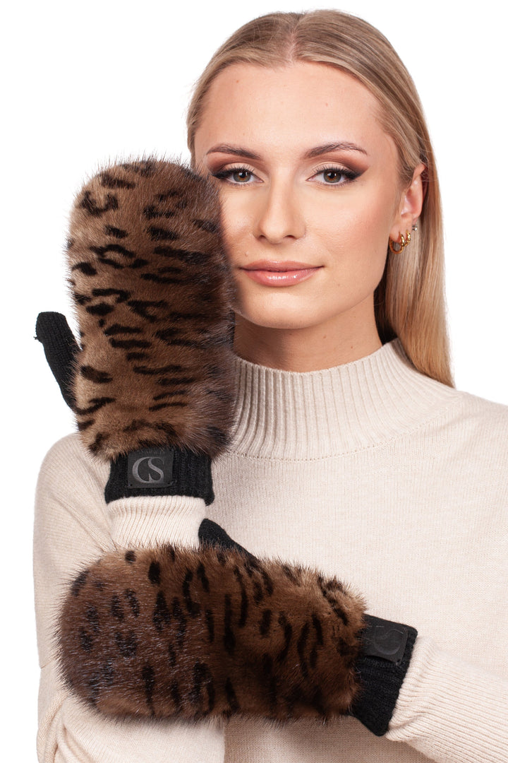 A woman with long blonde hair wears a beige sweater and FurbySD's Leopard Print Mink Fur Flip Top Mittens. She holds one to her cheek, smiling softly. These mittens, epitomizing ethical luxury, showcase mink fur accents and black cuffs.