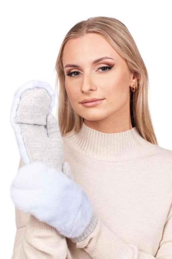 A person with long blonde hair is wearing a light beige turtleneck and holding up cozy Light Blue Mink Fur Flip Top Mittens by FurbySD. They are smiling softly against a plain white background, showcasing the perfect winter accessory.