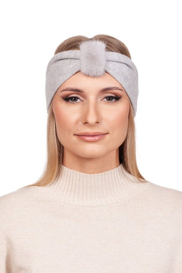 A person with long blond hair is wearing a FurbySD light grey cashmere and mink fur headband that features a pom-pom in the front. They are dressed in a beige turtleneck sweater, facing forward against a white background, capturing an understated luxury accessory feel.