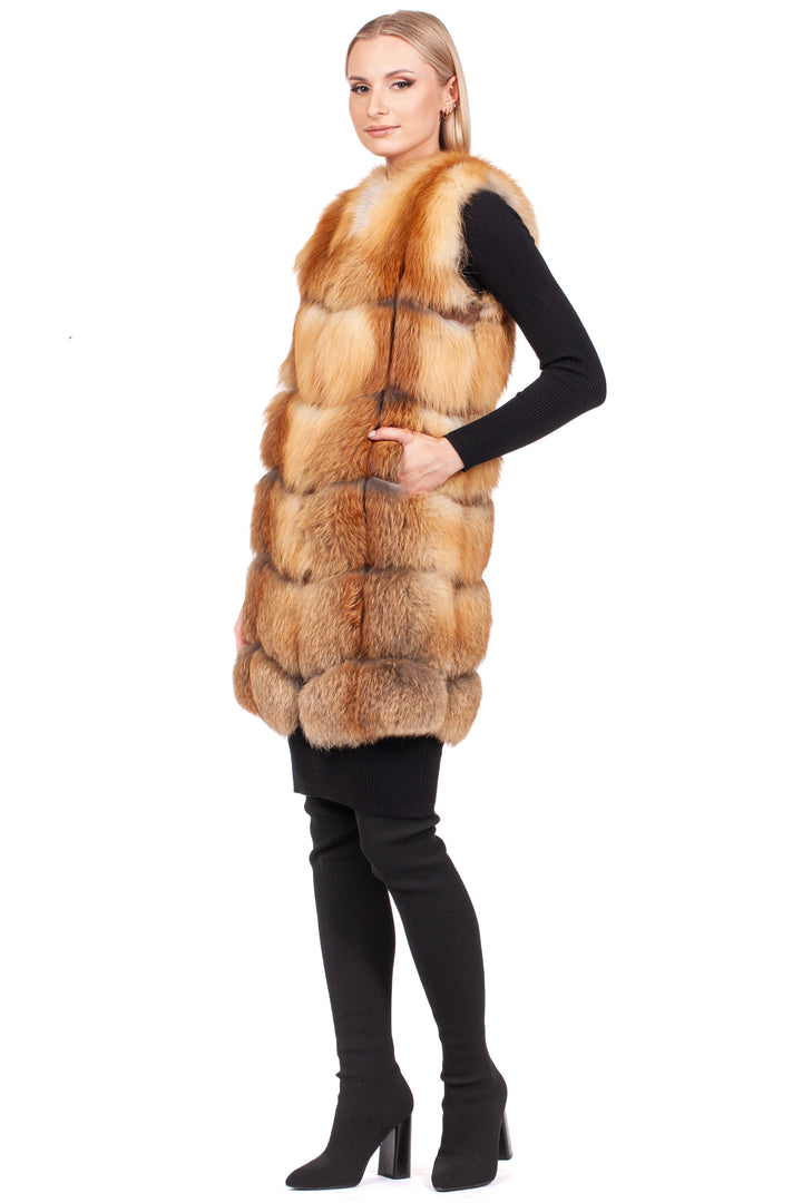 A person poses sideways in an elegant winter ensemble, featuring the Long Red Fox Fur Vest by FurbySD layered over a sleek black outfit with tall boots. Sporting short hair, they stand against a plain white background.