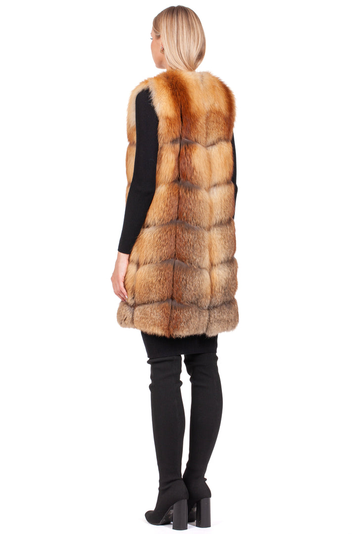 A model stands facing away, elegantly attired in a sleeveless Long Red Fox Fur Vest from FurbySD over a black top and pants—an essential piece of any sophisticated winter wardrobe. Her blonde hair cascades down, accompanied by high-heeled boots against a plain white backdrop.