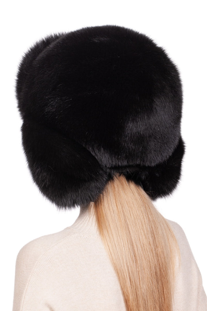 A person with long blonde hair wears a FurbySD Black Fox Fur Ushanka Trapper Hat, its large and fluffy design covering most of their head and ears, paired with a cream-colored sweater. This stylish winter accessory stands out against the plain white background.