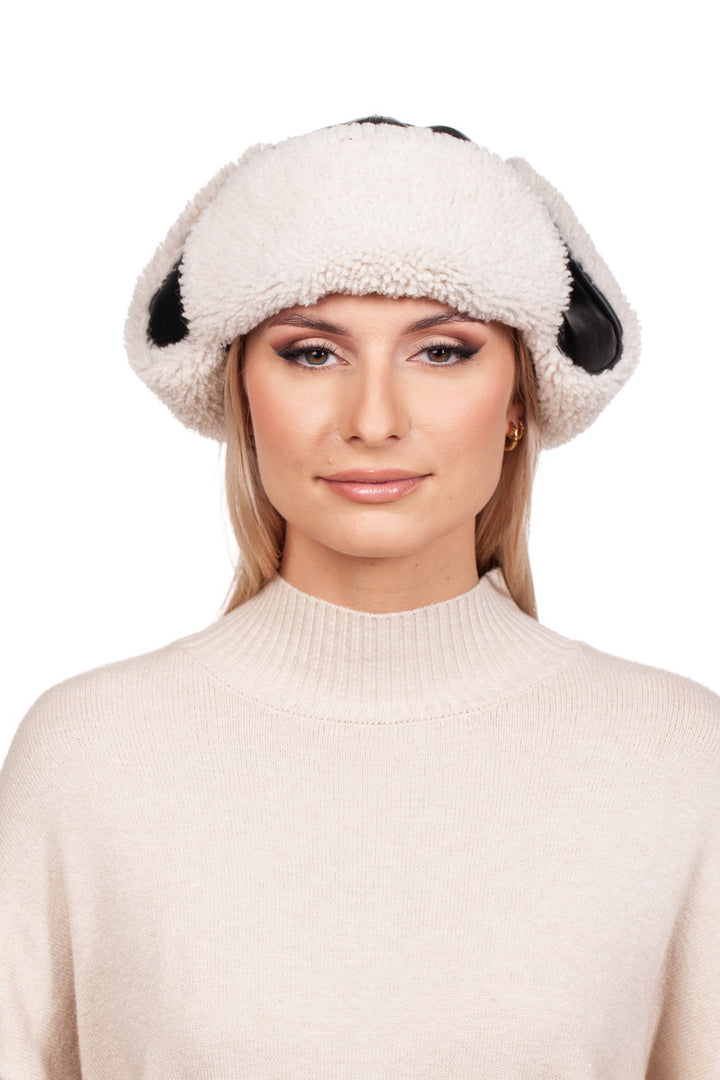 A person is wearing a whimsical FurbySD White Shearling Fur Aviator Bomber Hat adorned with playful black patches reminiscent of dog ears. Their long blonde hair complements a light-colored high-neck sweater, and they offer a slight smile against the plain white background, embodying chic simplicity.