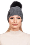 A person with long blonde hair is seen wearing the FurbySD Merino Beanie With Detachable Fox Bobble, featuring a hand-knitted gray design topped with a black pompom made of fox fur. They are styled in a cream-colored sweater and gaze directly at the camera against a white backdrop.