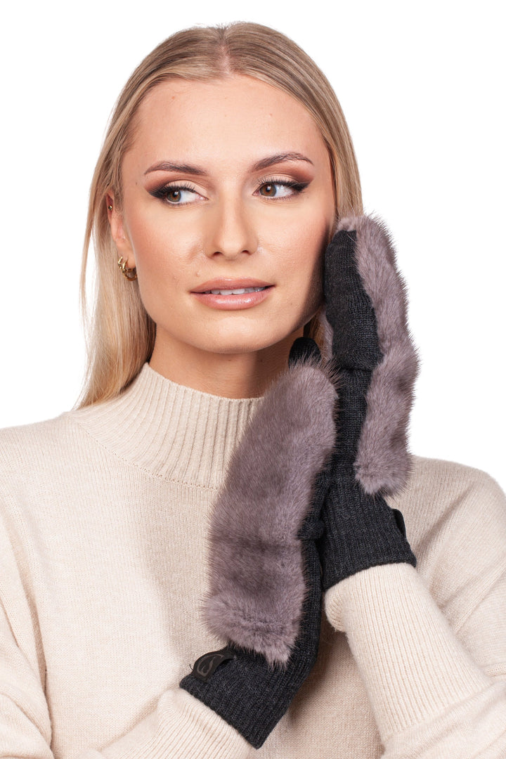 A person with long blonde hair is wearing a cream knit sweater and FurbySD's Silverblue Mink Fur Flip Top Mittens. They hold their gloved hands to one side of their face, looking to the side with a subtle smile.