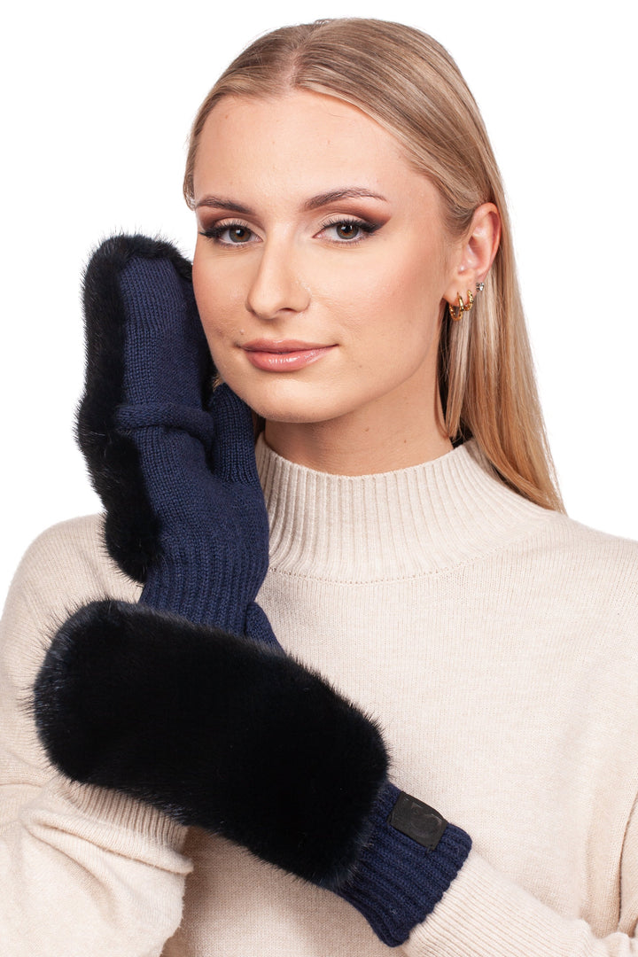 A person with long hair, dressed in a cream-colored turtleneck sweater, is holding a FurbySD Dark Blue Mink Fur Flip Top Mitten to their face, gazing directly at the camera.