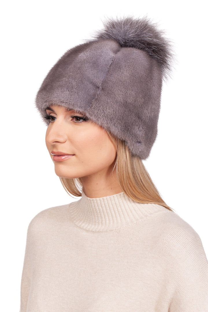 A person is wearing a Silver Blue Mink Fur Hat With Fox Pom Pom from FurbySD. They have long blonde hair and are dressed in a cream-colored sweater against a plain white background.