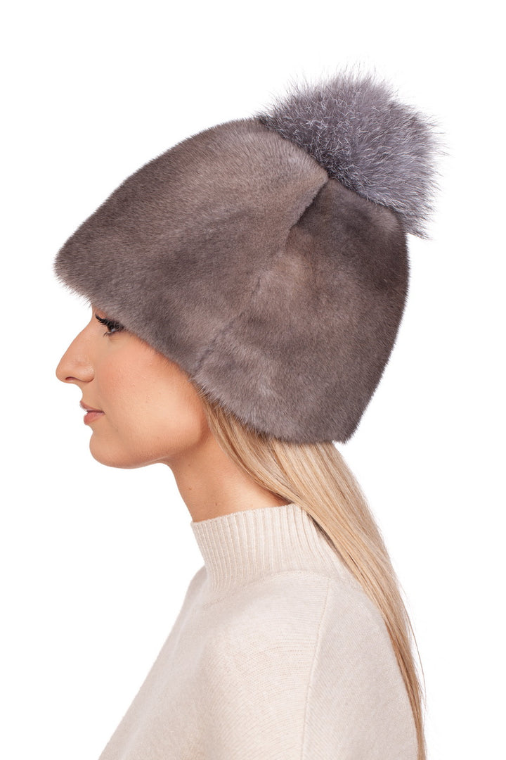 A side profile showcases the FurbySD Mink Fur Bobble Beanie Hat, featuring a plush gray design with a large fox fur pom-pom. The individual, whose long blonde hair cascades over a beige sweater, is positioned against a pristine white background.