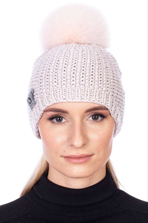 Woman wearing a pale pink merino wool handknit beanie with a matching fox fur pom-pom on a white background.