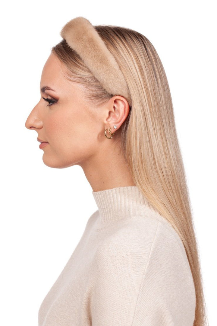 A woman with long blonde hair is wearing a FurbySD Beige Palomino mink fur headband paired with a matching turtleneck sweater. She showcases multiple earrings while posed in profile against a simple white background.