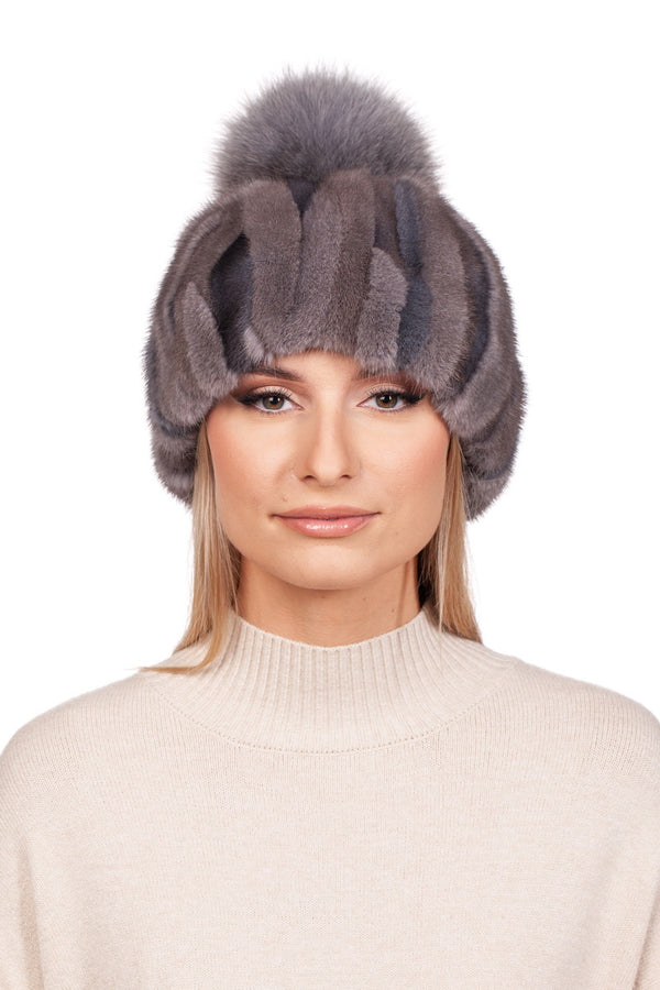 A person wearing a cozy Patched Grey Mink Fur Hat With Fox Bobble by FurbySD, paired with a cream-colored sweater, is set against a plain white background.