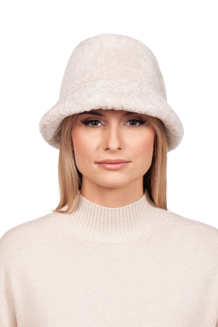 A person with long blonde hair is wearing a beige, textured Pearl White Astrakhan Fur Bucket Hat by FurbySD and a matching beige sweater with a high neckline. They have a neutral facial expression and are looking directly at the camera against a plain white background.