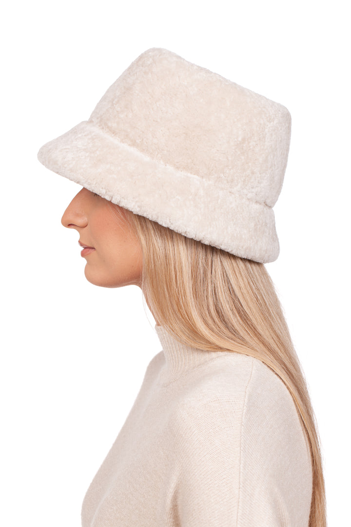 Side profile of a woman with long blonde hair, wearing a FurbySD Pearl White Astrakhan Fur Bucket Hat and a matching cream turtleneck sweater. The background is plain white.