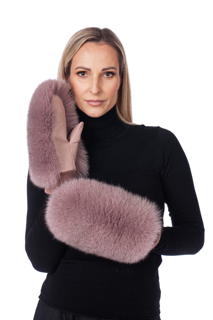 A woman with long blonde hair stands against a plain white background, modeling the Pink Fox Fur Collar And Mittens Set by FurbySD. Dressed in a black turtleneck sweater, she holds one fluffy mauve mitten up by her face, displaying both the front and back of the mittens. Her elegant look is complemented by the pink fox fur collar.