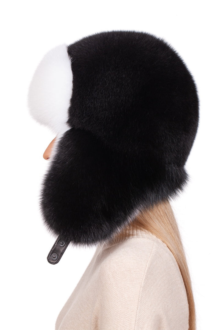 A person wearing a luxurious Fox Fur Ushanka Trapper Hat from FurbySD, featuring a fluffy black and white design with ear flaps, stands against a white background. The hat's soft texture is highlighted as the individual dons a beige top.