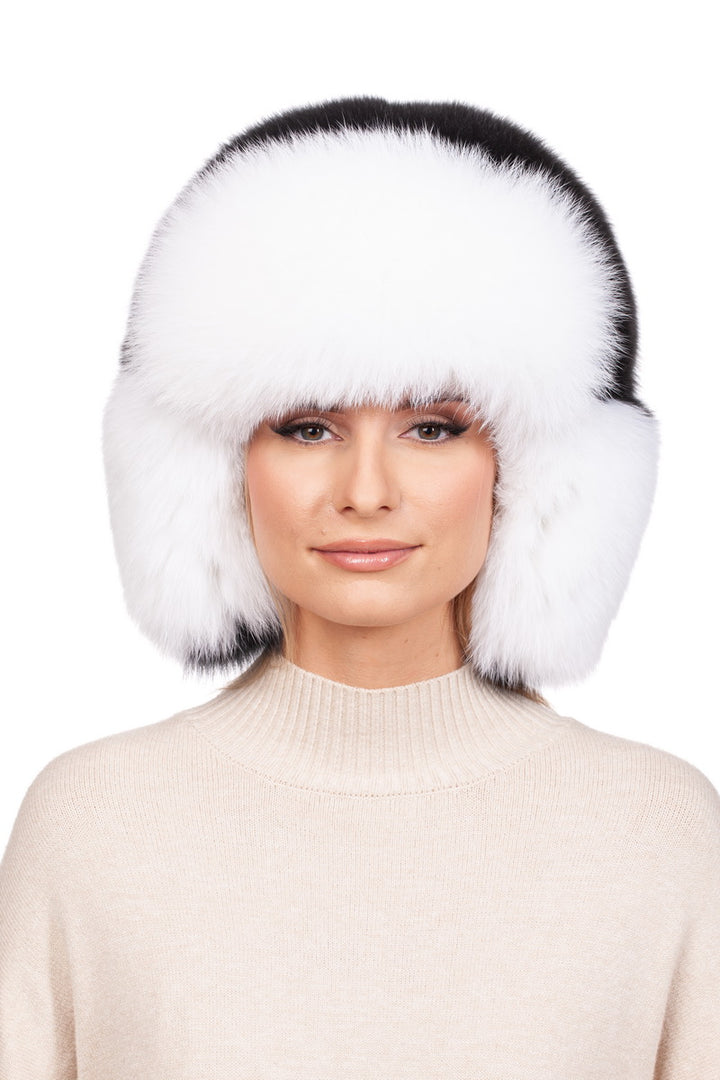 A person is wearing the FurbySD Fox Fur Ushanka Trapper Hat, a large, fluffy accessory in white and black fur with ear flaps. They have a neutral expression and are dressed in a cream-colored knit sweater, standing against a white background that emphasizes this essential winter accessory.