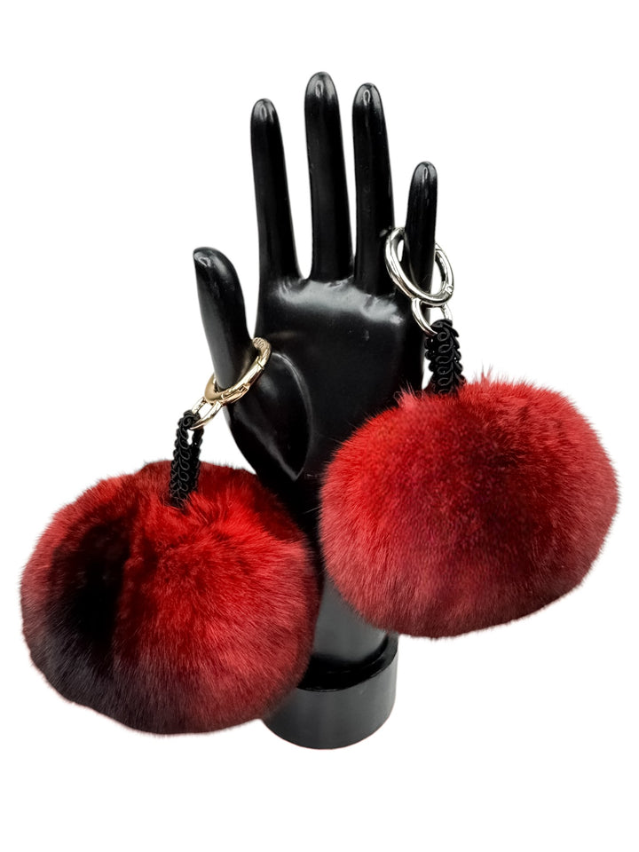 A sleek black mannequin hand showcases two opulent Red Chinchilla Fur Bag Charms by FurbySD, featuring metal rings. Perfect for embellishing a bag, these luxurious accessories elevate any look with a touch of elegance.