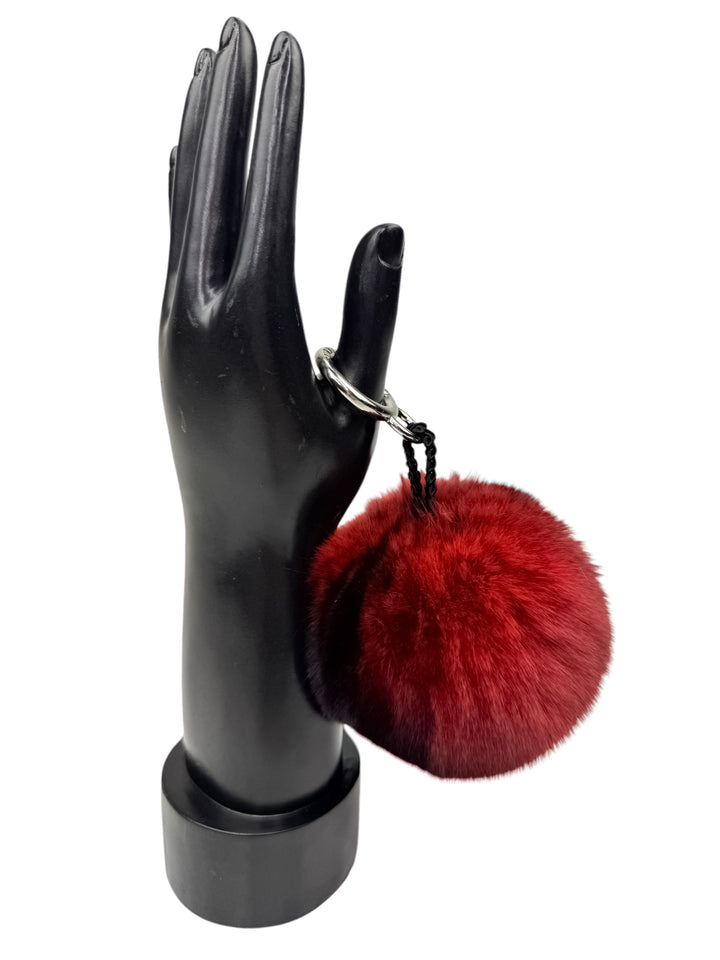 A black mannequin hand elegantly displays the Red Chinchilla Fur Bag Charm by FurbySD, a luxurious red, fluffy pom-pom keychain attached to the ring finger. This exquisite accessory adds a pop of color to the minimalist setup, making it a stunning addition to any bag.