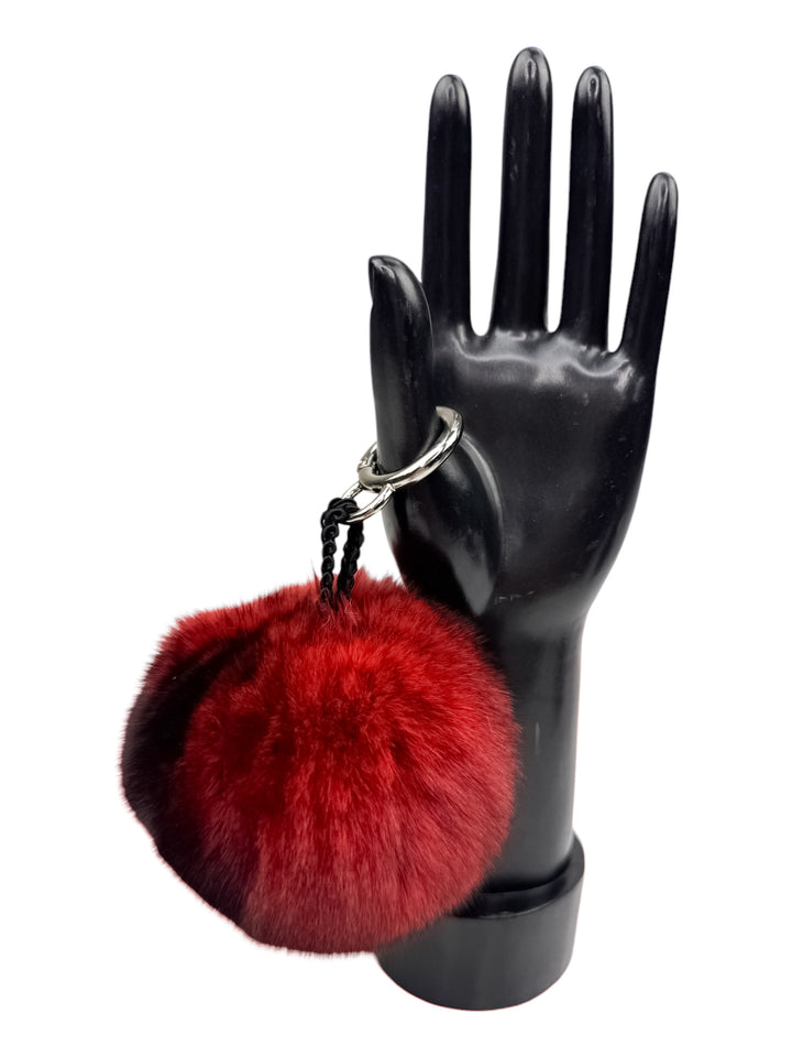 A black mannequin hand elegantly displays the Red Chinchilla Fur Bag Charm by FurbySD, a true luxury accessory. The bag charm, soft and spherical, is attached with a silver ring and contrasts beautifully with the smooth, dark finish of the hand display.