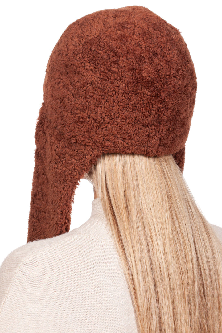 A person with long, blonde hair is wearing the Rusty Brown Shearling Fur Earflap Hat by FurbySD. Seen from the back, this winter accessory in a textured rusty brown shade covers most of their back and gracefully complements a light-colored sweater.