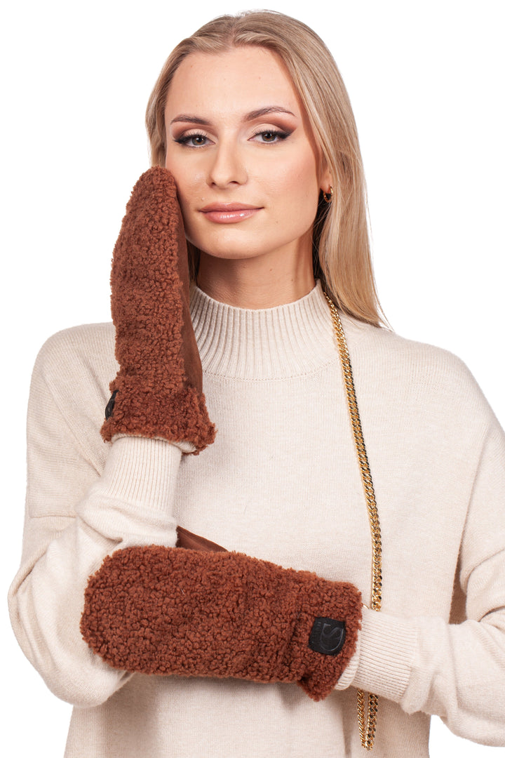 A woman with long blonde hair is wearing a white sweater and FurbySD's Brown Shearling Fur Mittens With Detachable Chain. With a gentle expression, she holds one mitten to her face against the plain white background.