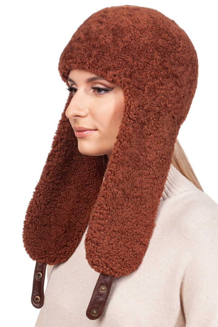 A person wearing a Rusty Brown Shearling Fur Earflap Hat from FurbySD with buttoned leather straps. They have long blonde hair and are dressed in a beige sweater. This hat, an ideal winter accessory, covers their ears, offering both warmth and style.