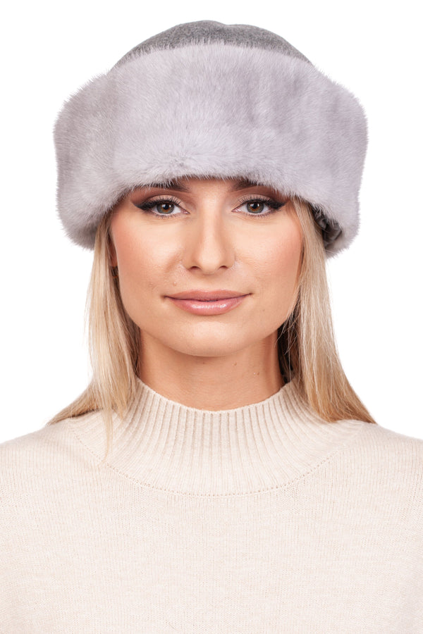 A person with long blonde hair wears the Sapphire Mink Fur and Cashmere Hat by FurbySD along with a cream-colored knitted turtleneck sweater. The background is white, highlighting their serene expression and showcasing the hat as an ideal winter accessory.