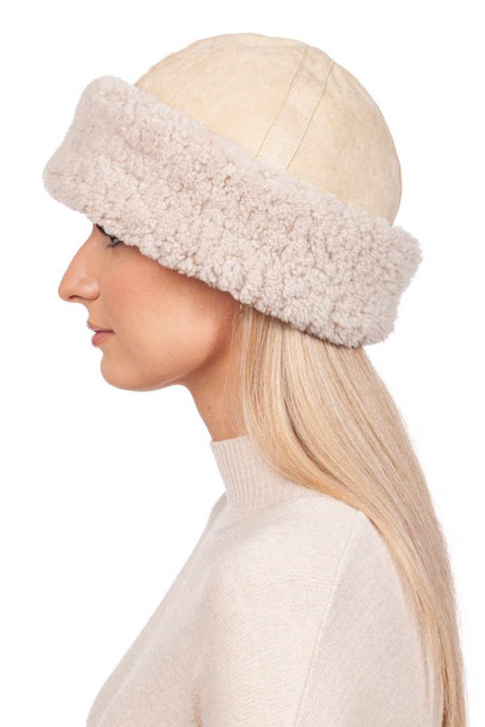 A person with long blonde hair is wearing a Beige Shearling Fur Roller Hat by FurbySD, which serves as a luxury winter accessory. Dressed in a cream-colored sweater, they face left against a plain white background.