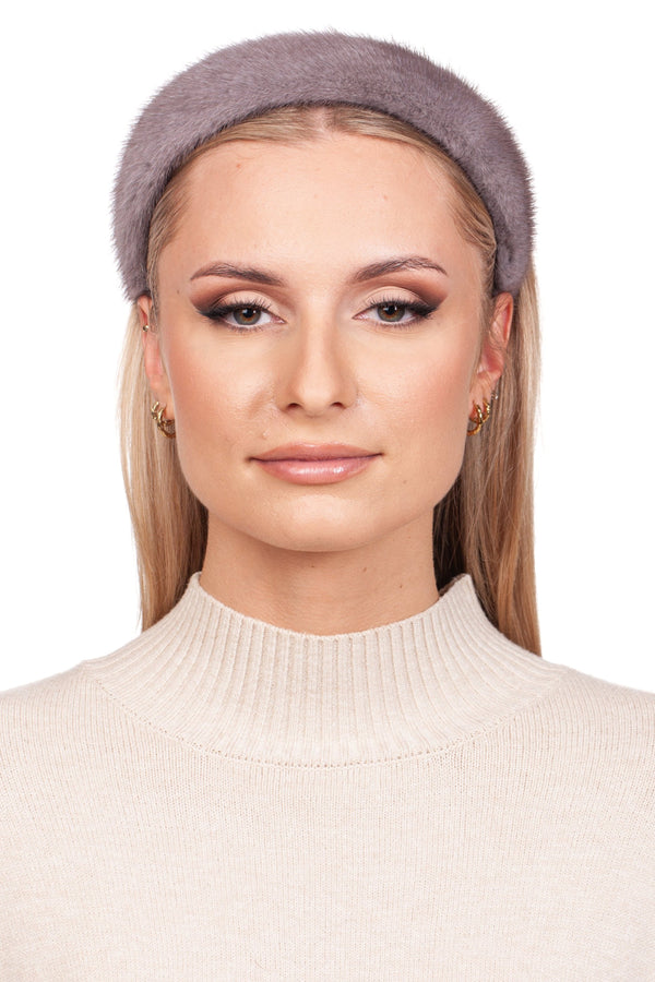 A person wears a beige ribbed turtleneck and the Grey Mink Fur Headband from FurbySD, exuding elegance with their long blonde hair, subtle makeup, and gold hoop earrings. They pose against a plain white background, showcasing the charm of luxury winter accessories.